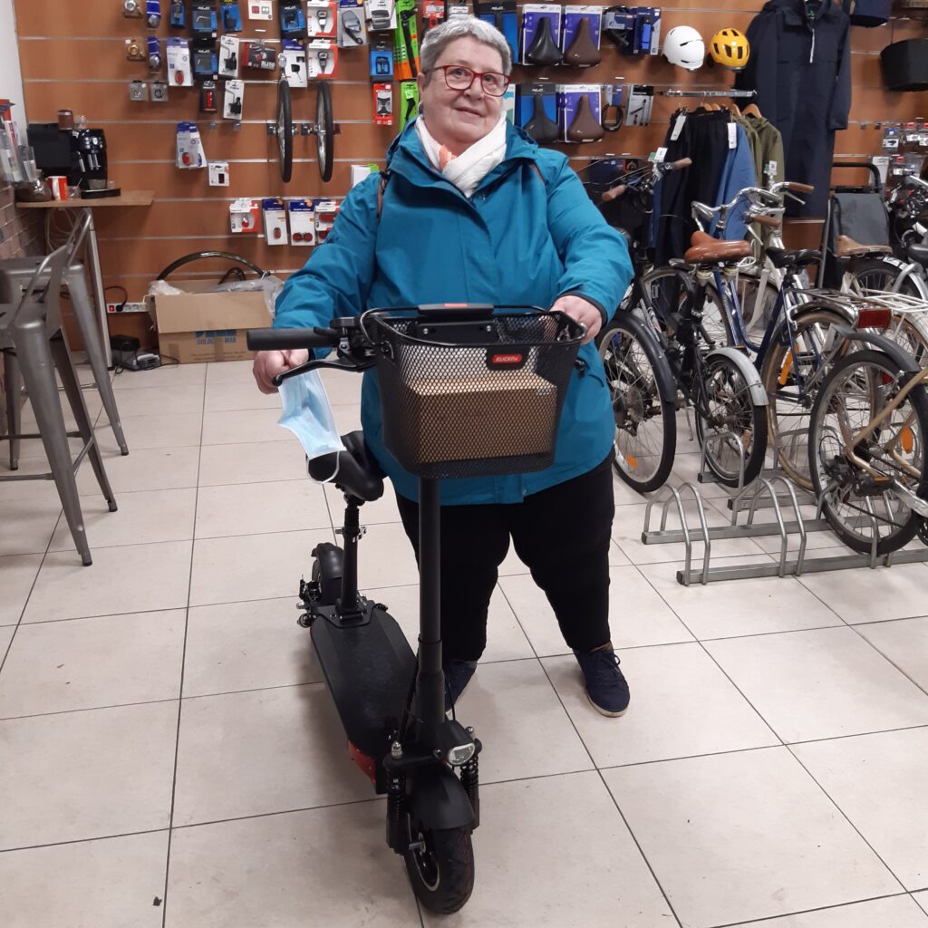 femme senior avec sa trottinette électrique équipée d un panier de transport à l' avant, dans le magasin Citytrott rempli d'accessoires casques, selles en tout genre et vélos électriques