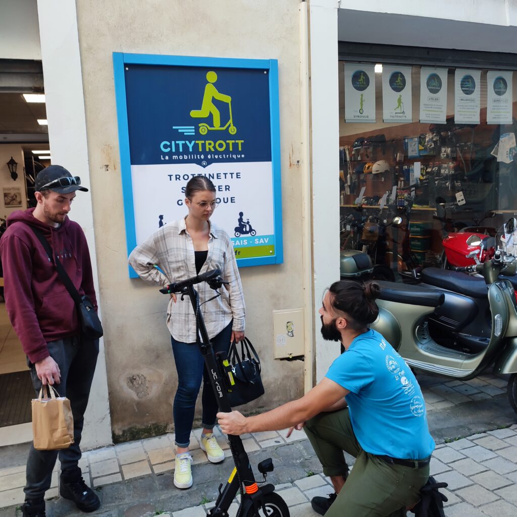 Devant un magasin Citytrott , deux jeunes acheteurs profitent des conseils d'un vendeur d'une trottinette électrique. A arrière plan deux scooters électriques