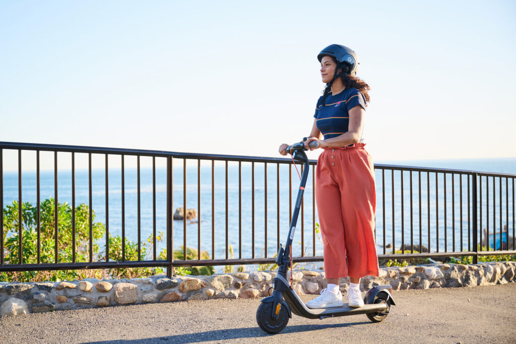Une jeune femme en pantalon rouge er baskets se promène le long de la mer à trottinette électrique