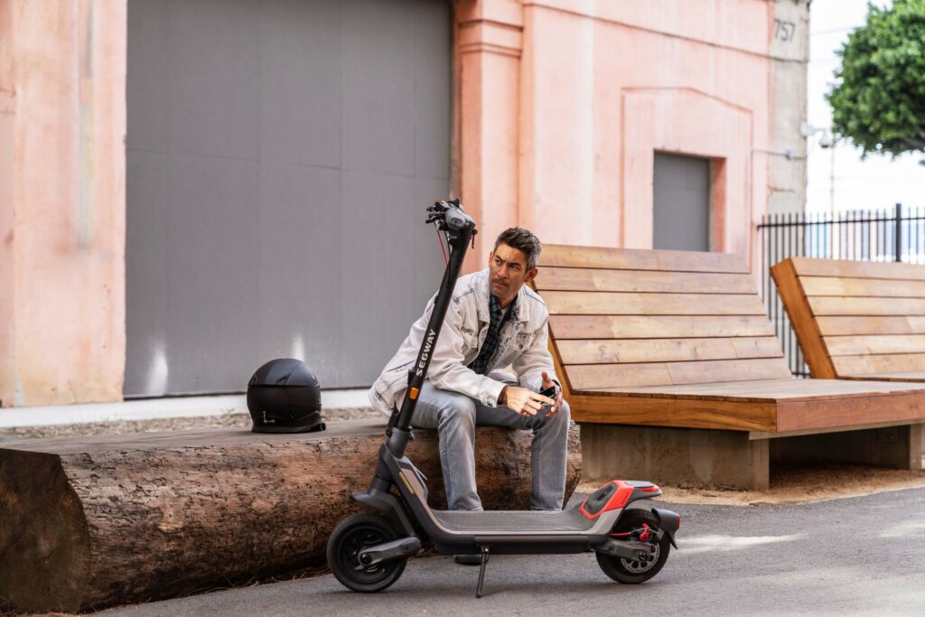 Un homme avec une veste en jean est assis sur un banc avec un casque posé à côté de lui et en face de sa trottinette électrique Segway un modèle sportif, puissant et confortable