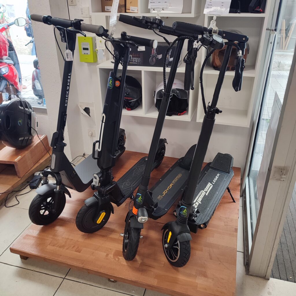 présentoir de 4 trottinettes électriques de différentes marques à différents prix et présentation à l'arrière d'un choix de casques blanc, marron, noir